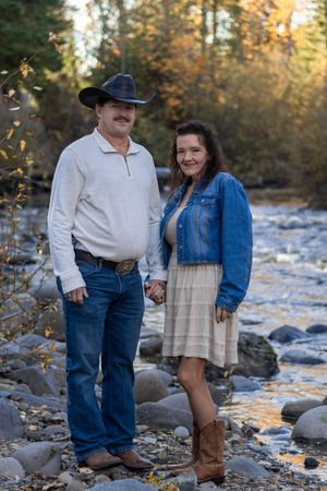 Couple holding hands by rocky creek in autumn, man in black cowboy hat, woman in denim jacket and boots.