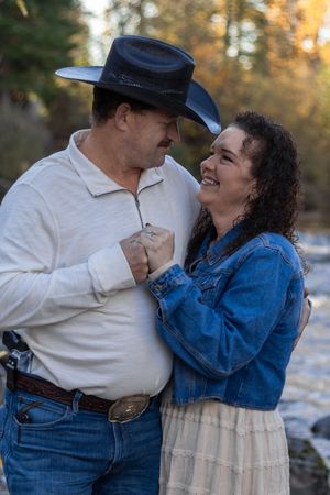 Laughing couple embracing by autumn creek, man in black cowboy hat and white shirt, woman in denim jacket.