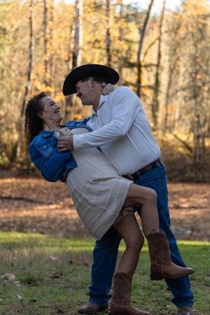 Man in cowboy hat lifting laughing woman in denim jacket during outdoor fall engagement photo session.