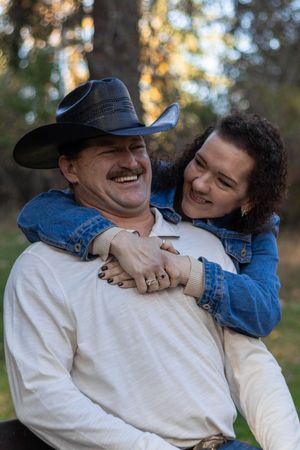 Woman in denim jacket hugging man in cowboy hat from behind, both laughing joyfully outdoors.