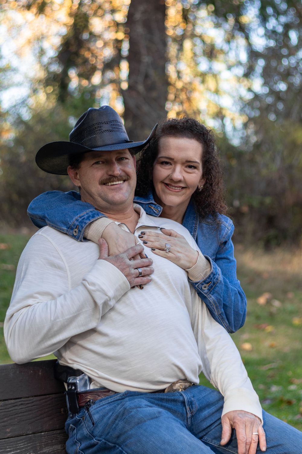 Smiling couple posing on bench in autumn park, man in cowboy hat, woman in denim jacket embracing him.
