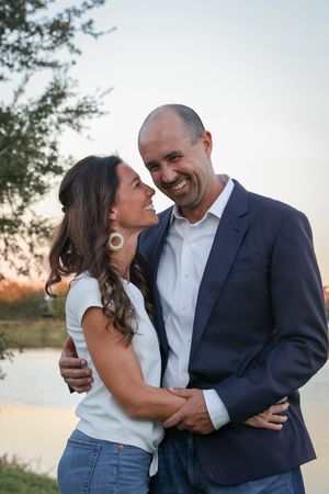 A couple embraces and smiles at each other outdoors during golden hour by a lake.