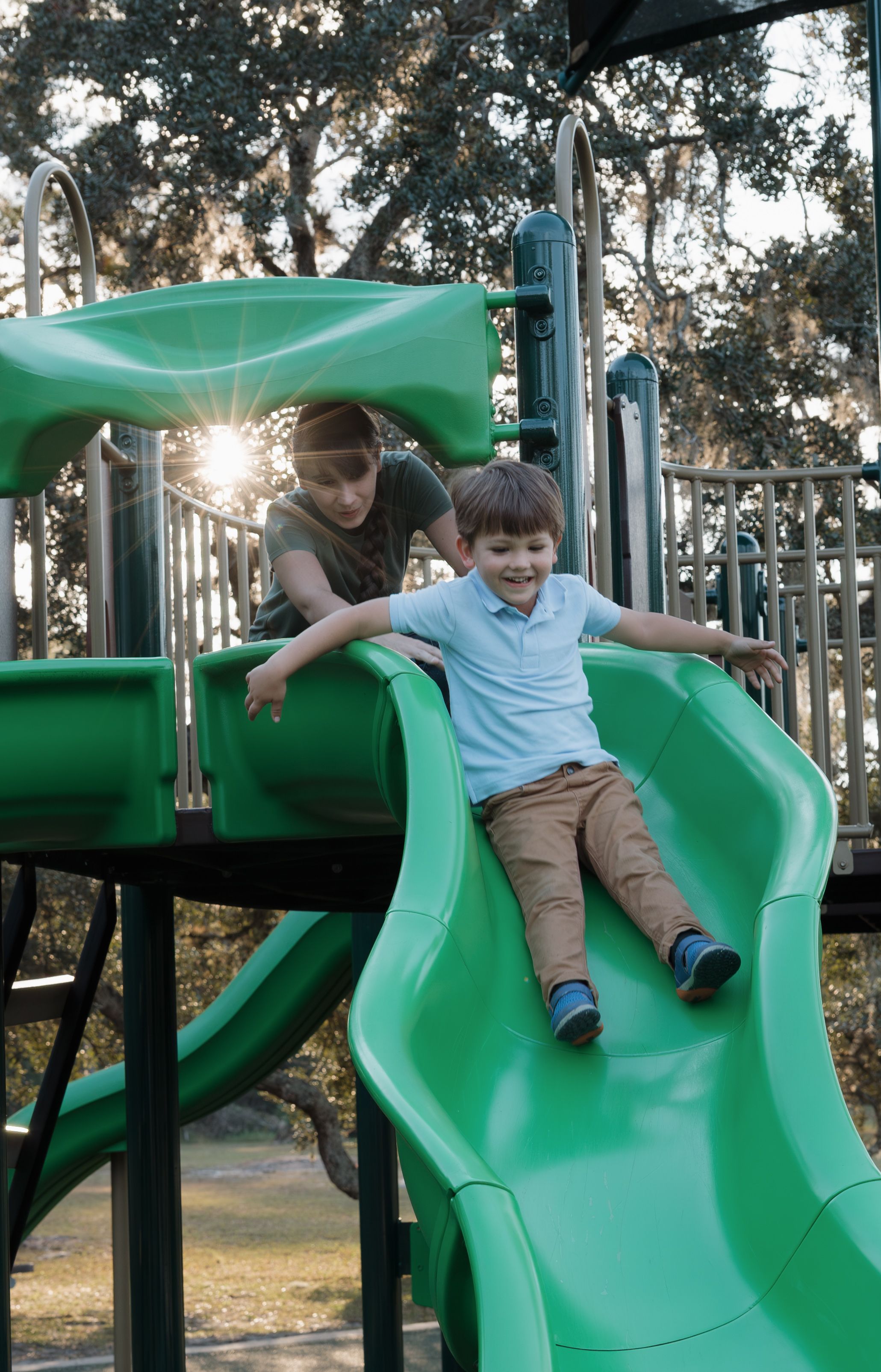 Child playing and sliding on green playground equipment at sunset.
