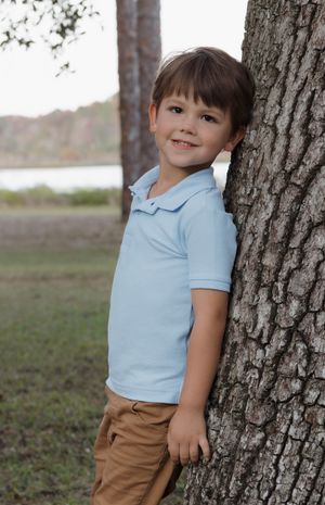 Child in light blue shirt and khaki pants posing by large tree trunk.