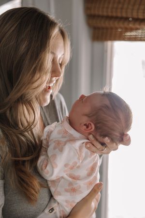 Mother holding newborn baby in white floral outfit by window.