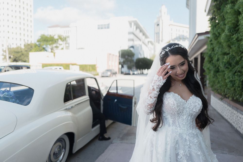 Bride in white lace wedding gown and tiara stands beside a classic white car, smiling on her wedding day.