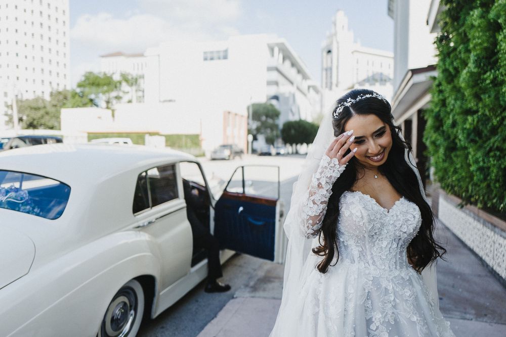 A bride in an elegant white lace wedding gown poses near a vintage white car on a sunny day.