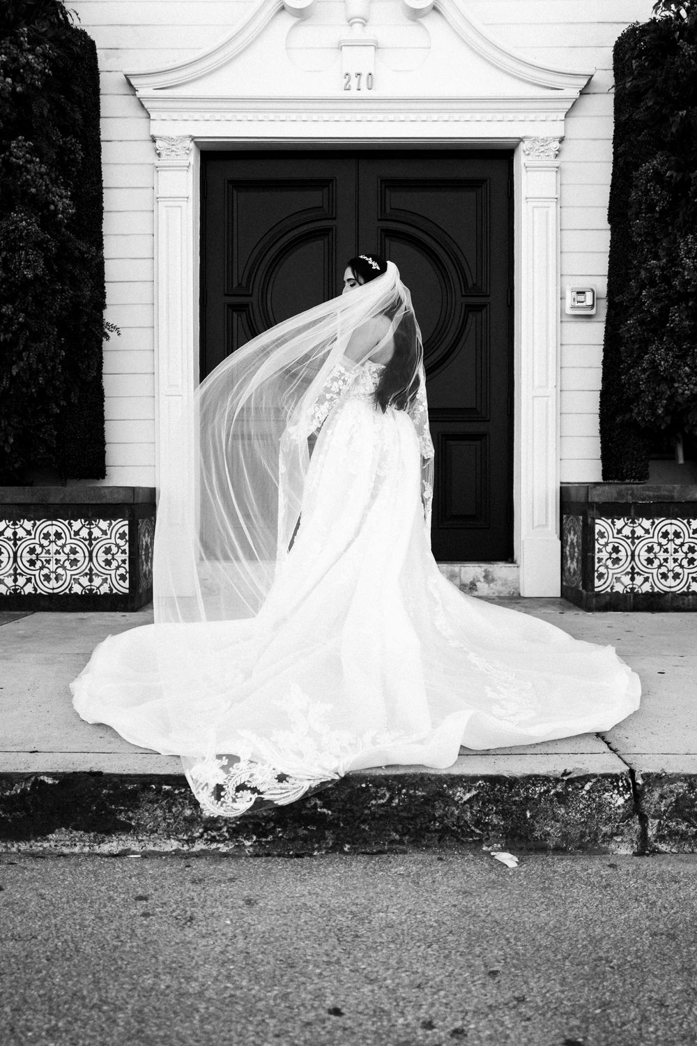 A flowing white wedding dress billows dramatically in the wind against a classic white building facade.