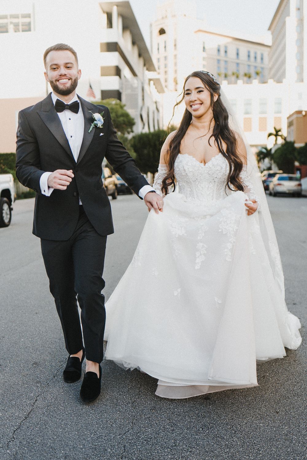 Newlywed couple holding hands and walking together on an urban street in formal wedding attire.