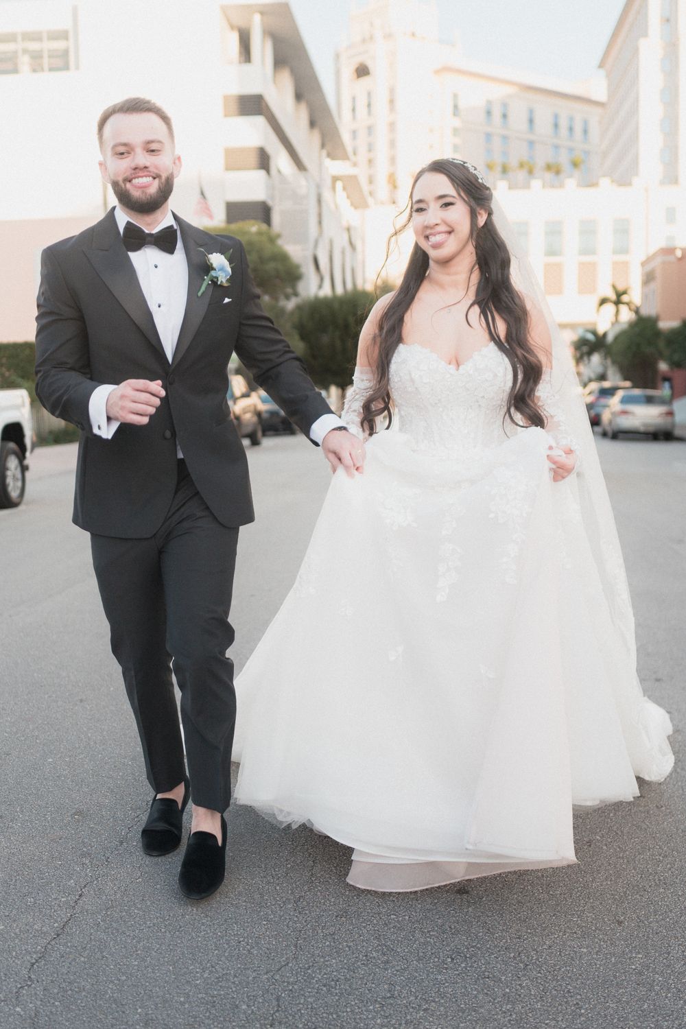Bride in white lace gown and groom in black tuxedo walk hand in hand on city street on their wedding day.