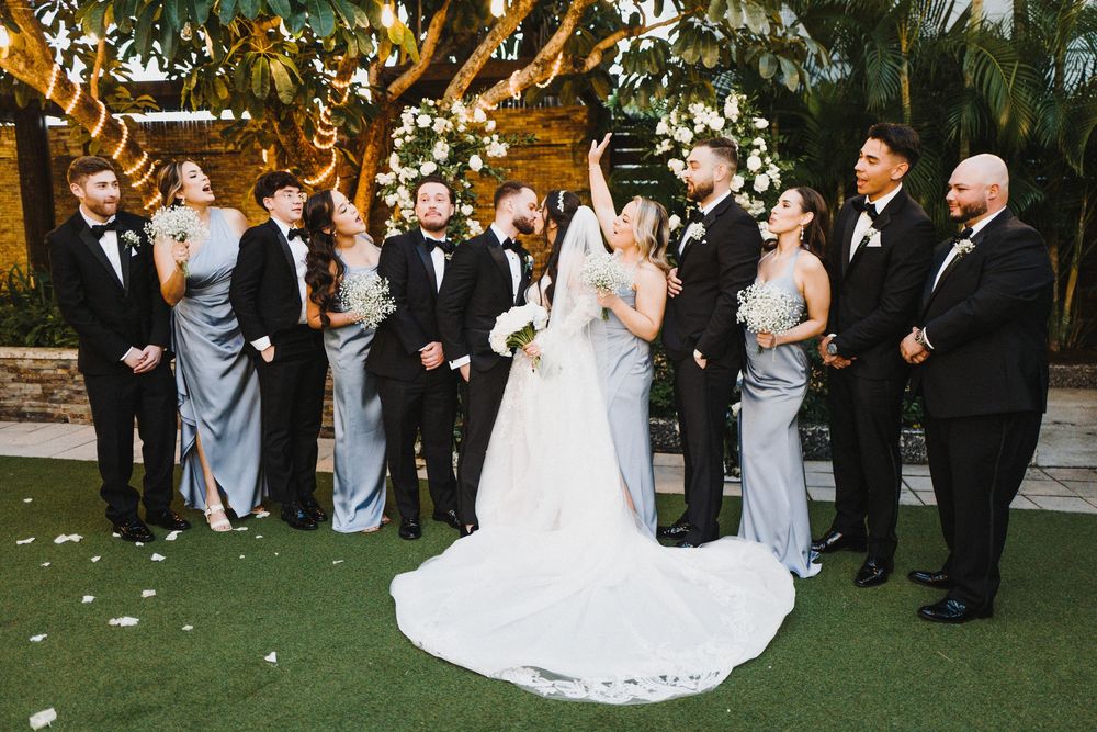 Wedding party in formal attire celebrates outdoors under palm trees during golden hour reception.