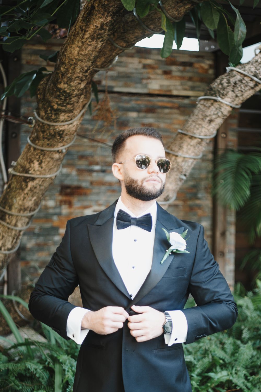 A groom in a classic black tuxedo and bowtie poses in front of a stone wall and tropical greenery.