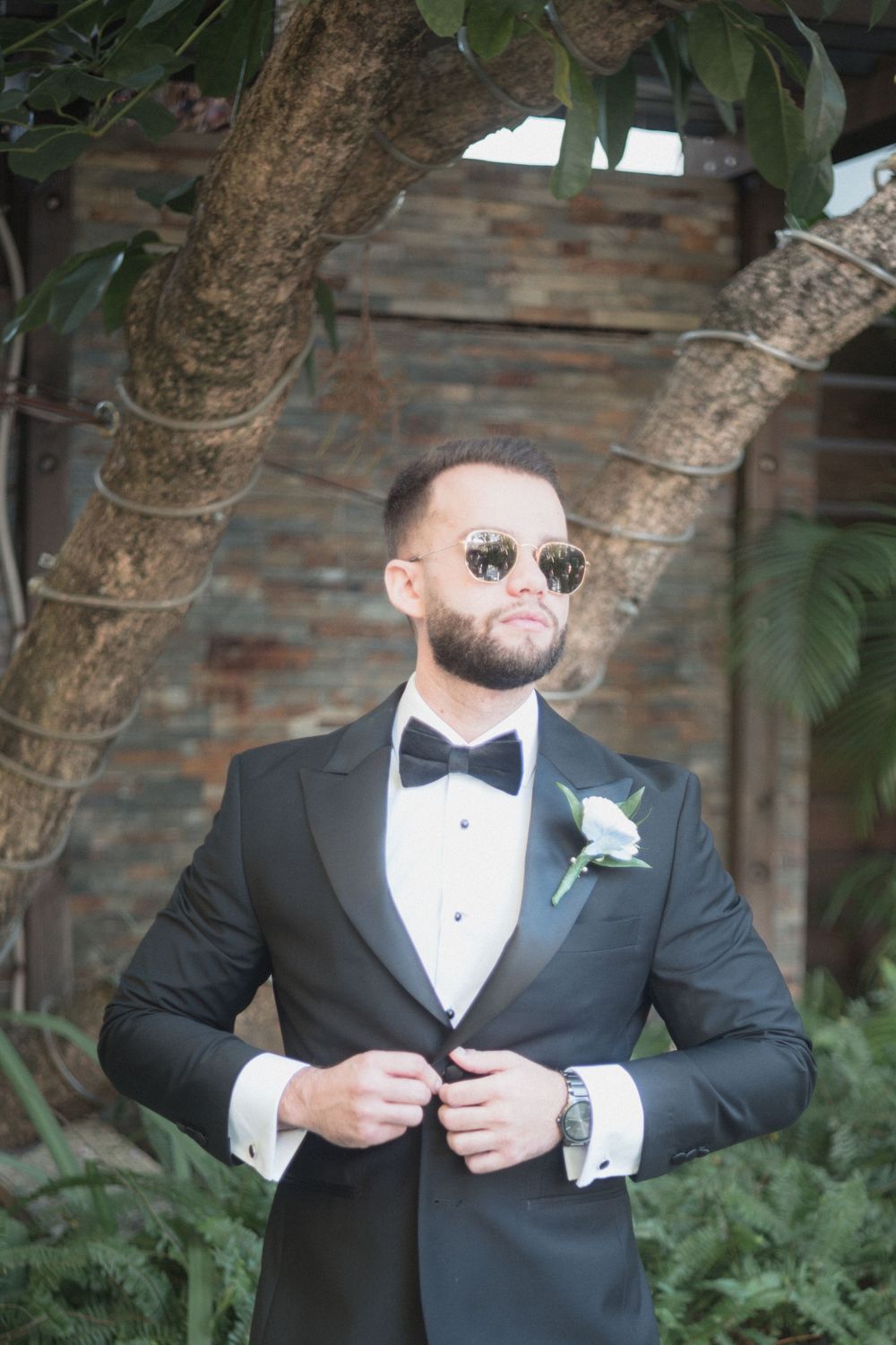 Groom in black tuxedo with bow tie and sunglasses, adjusting jacket with floral boutonniere outdoors.