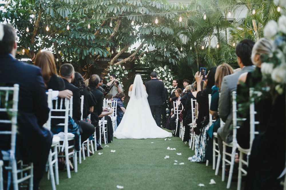 Elegant outdoor wedding ceremony setup with white chairs lined up under trees and white floral decorations.