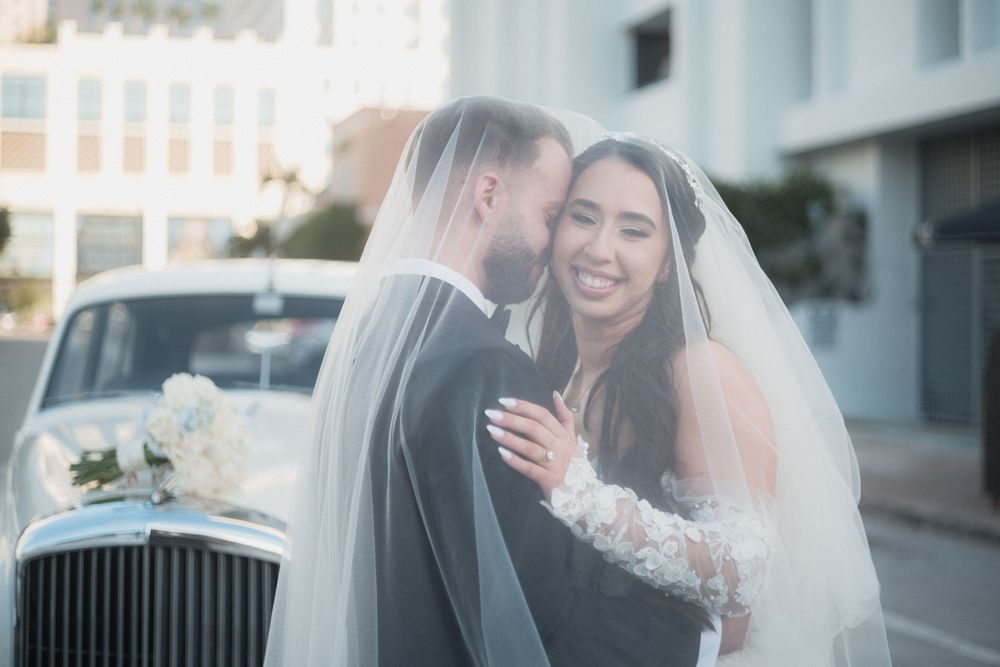 Bride and groom share romantic moment under veil beside vintage white car on wedding day.