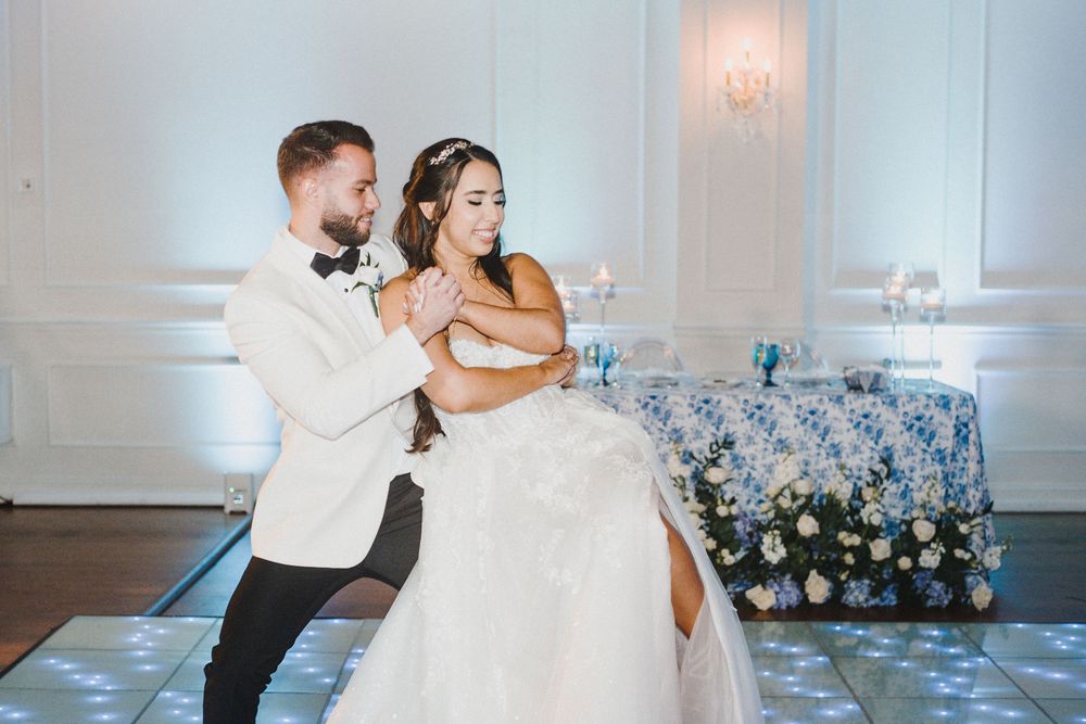 A couple shares a romantic dance in an elegant white ballroom decorated with floral arrangements and soft lighting.