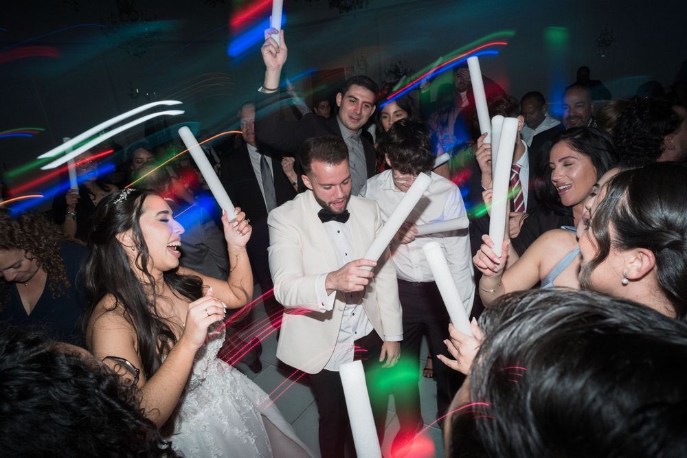 Wedding reception dance floor celebration with guests waving colorful glow sticks, bride in white gown and groom in white tuxedo.