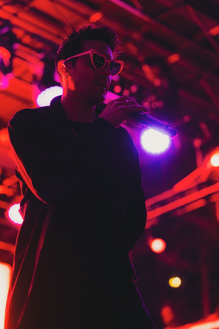 A silhouetted performer in sunglasses sings on stage illuminated by colorful concert lighting.