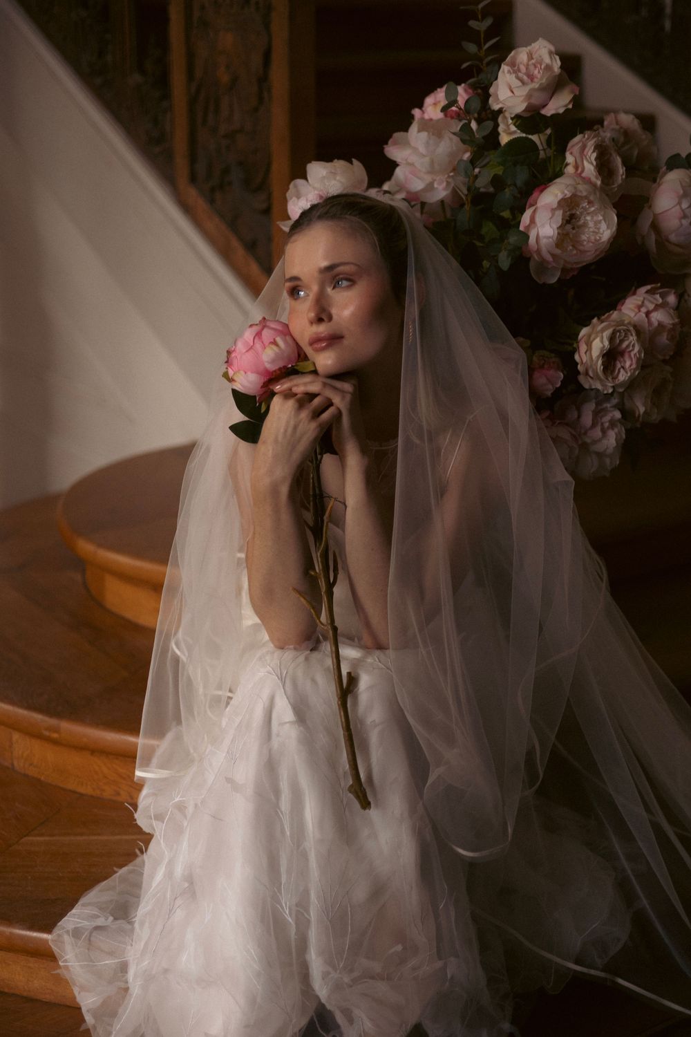 A bride in a white wedding dress and veil poses on stairs with pink roses in a moody, romantic setting.