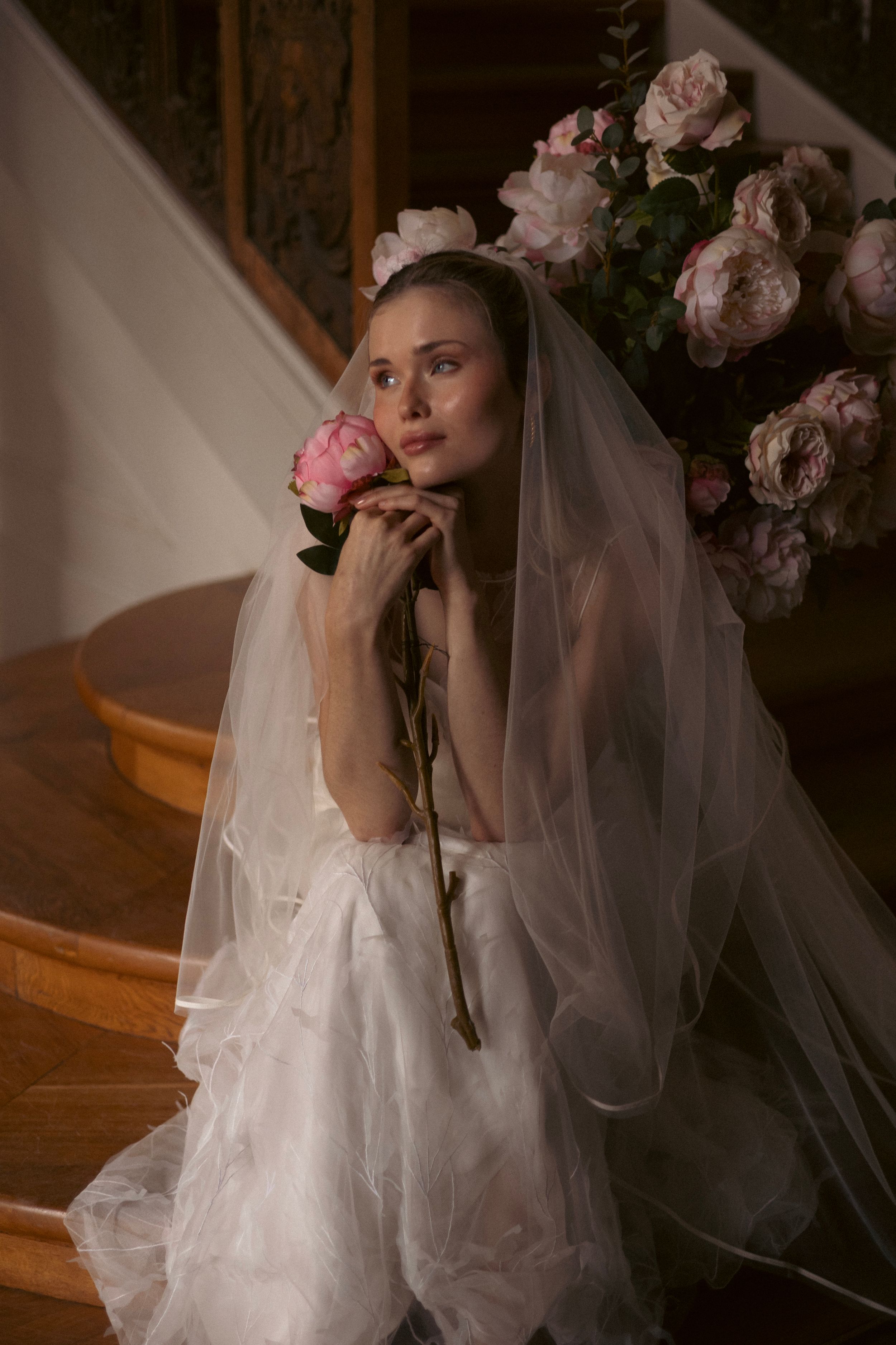 A bride in a white wedding dress and veil poses on stairs with pink roses in a moody, romantic setting.