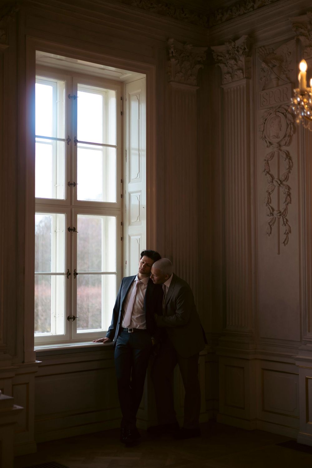 A couple stands in silhouette by a tall vintage window in an ornate room with decorative molding and a chandelier.