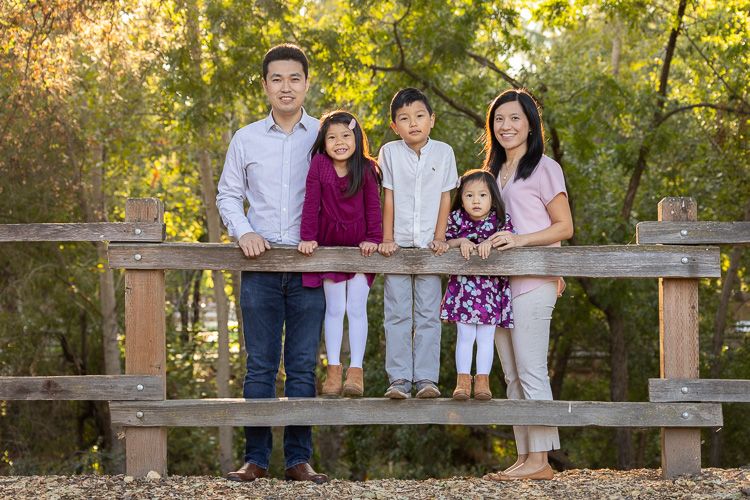 A family stands together on a wooden fence in a park during golden hour with autumn trees in the background.