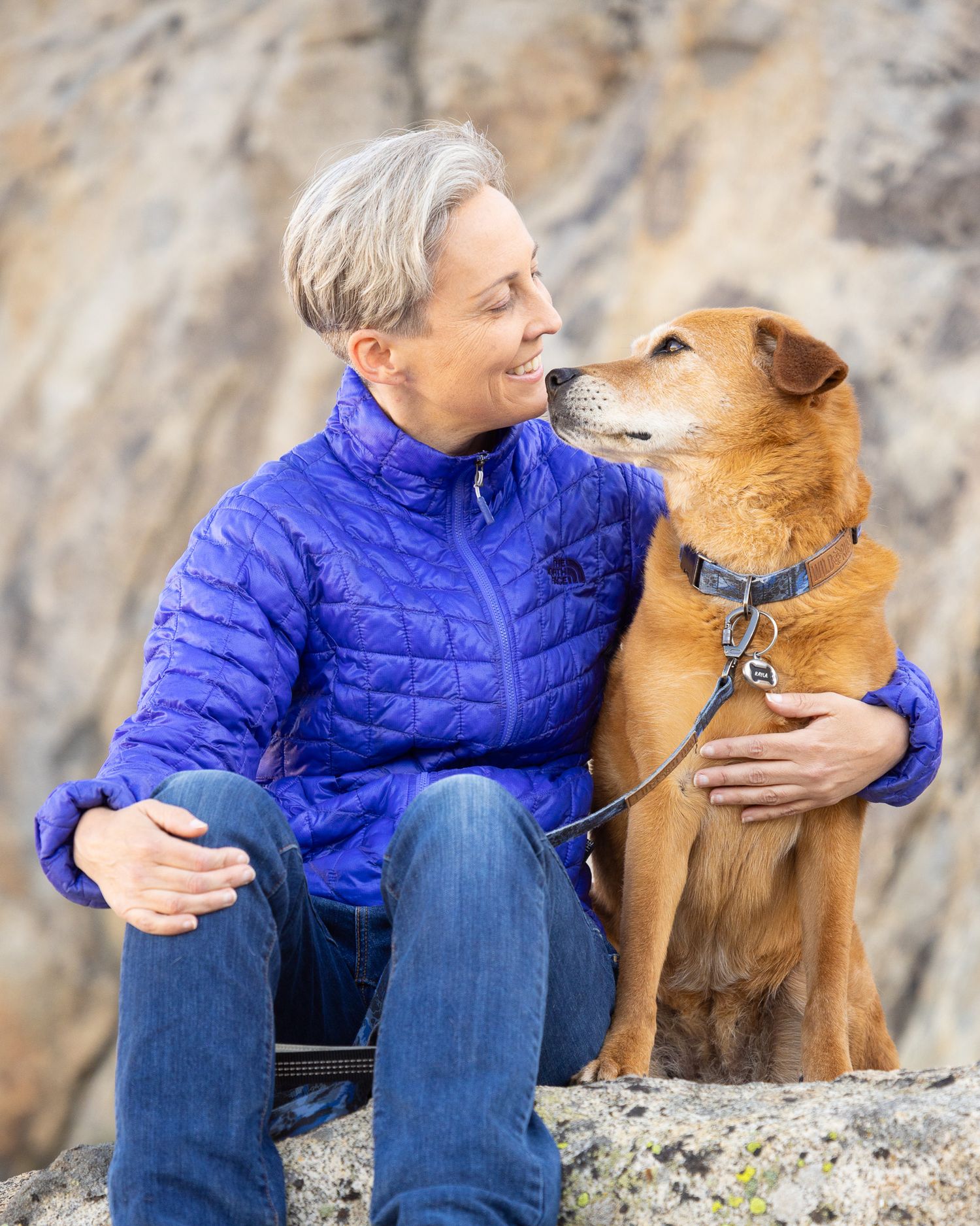 A lady shares a tender moment with her brown dog during a family photo session, while sitting outdoors on a rock