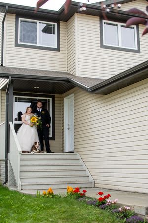 Wedding party poses on front porch steps of beige house with sunflowers and red tulips in the garden.
