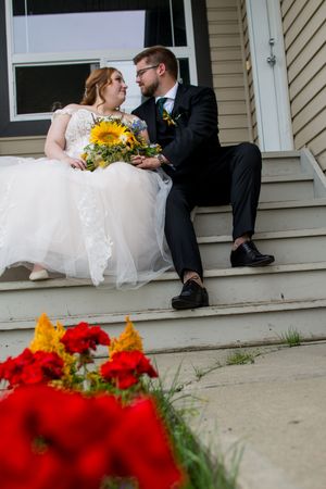 Newlyweds sit on porch steps with bright red flowers in the foreground and sunflowers in the bride's bouquet.