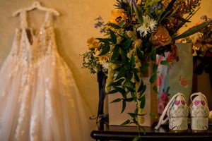 Wedding dress hanging beside floral arrangements and bridal shoes against a golden wall.