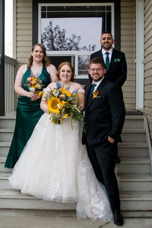 Wedding couple and attendants in black and emerald attire stand on white porch with sunflower bouquets.
