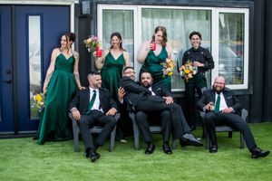 Wedding party lounges on grass wearing emerald green dresses and black suits against blue building.