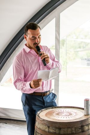 Person in pink button-down shirt and blue pants giving speech while leaning on wooden barrel.