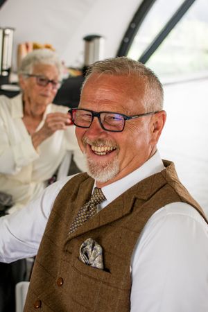 A smiling guest in a brown tweed vest and glasses enjoys a wedding celebration.