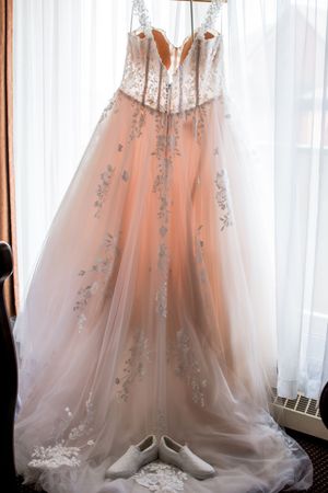 Elegant blush wedding dress with intricate beading and flowing tulle skirt hanging against white curtains.
