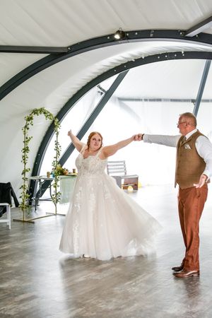 A couple performs a playful dance move in a modern venue with arched ceiling and greenery decor.