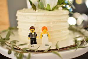 White wedding cake decorated with greenery and custom LEGO bride and groom cake toppers.