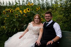 Wedding couple sits on bench in front of tall blooming sunflower garden.