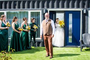 Wedding party poses outdoors against blue shipping containers and green grass on a sunny day.