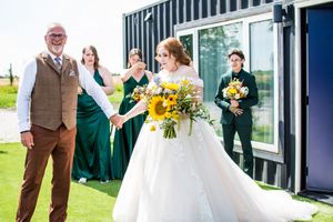 Wedding party in emerald green and brown attire posing outdoors with sunflowers.