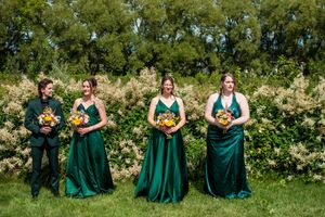 Wedding party in emerald green dresses poses together on a sunny grassy hillside.