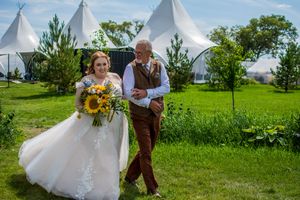 Couple in wedding attire dances and twirls in a green field with white teepee tents in the background.