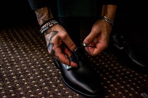 Close-up of hands with jewelry and tattoos tying formal black dress shoes on a patterned carpet.