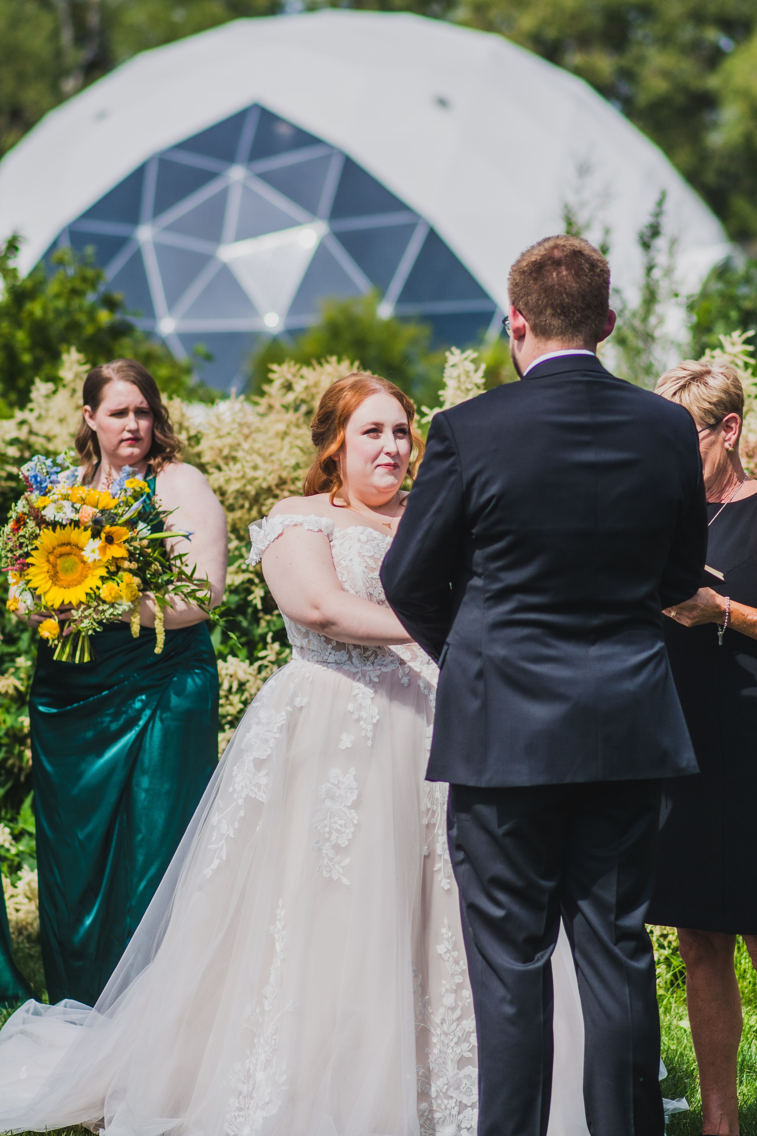 Wedding ceremony in front of a modern geodesic dome structure with sunflowers and emerald green accents.