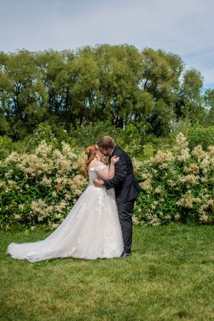 Romantic wedding couple embraces on grassy lawn with blooming white flowers and trees in background.
