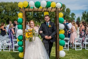 Outdoor wedding ceremony setup with white and green balloon arch and white chairs on grass.