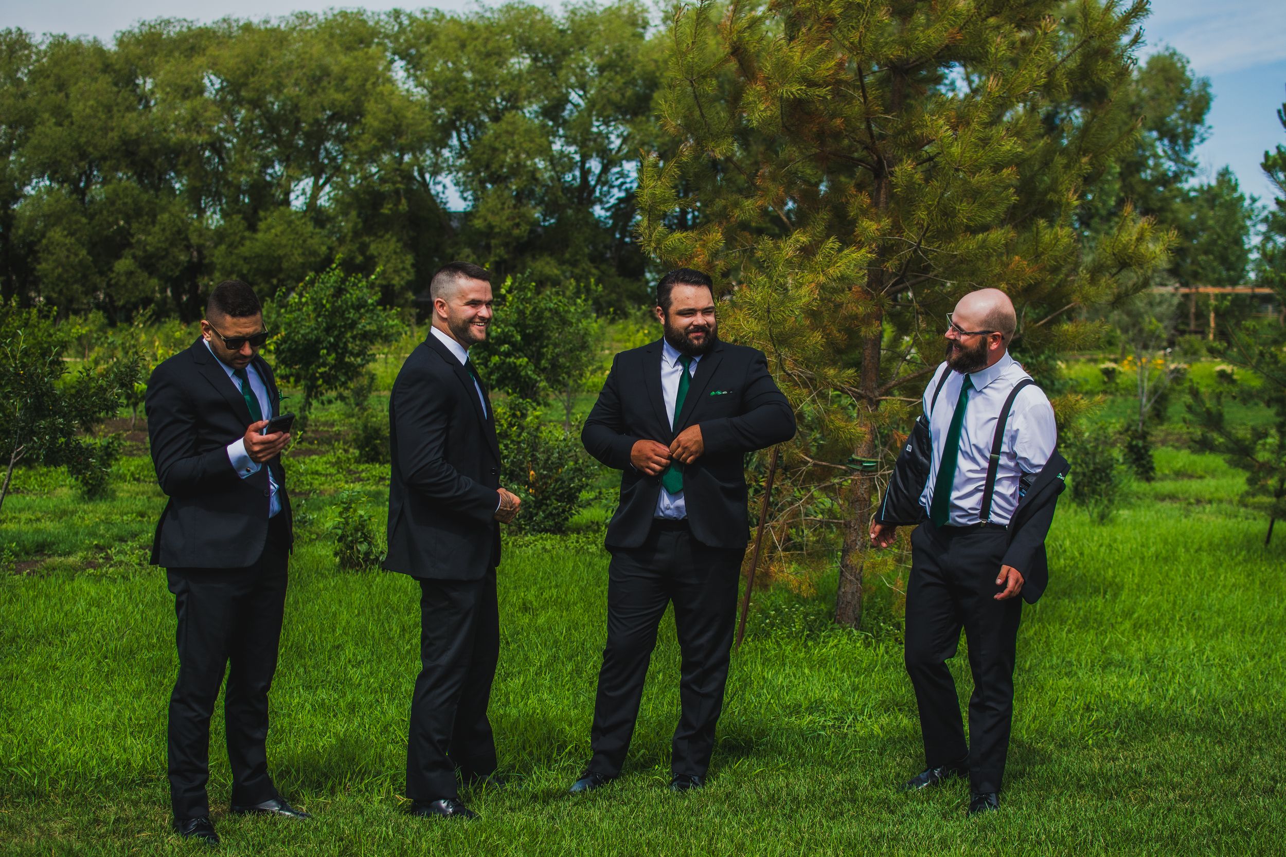 Wedding party members stand in a line on lush green grass with pine trees in the background.