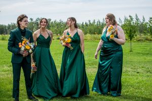 Bridesmaids in elegant emerald green dresses stand together on grassy field during outdoor wedding.