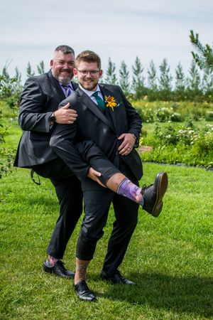 Groomsmen show off colorful socks while kneeling on green grass in formal black suits.