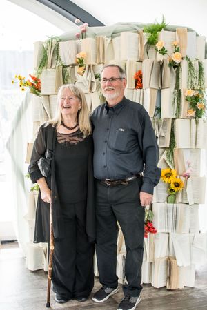 Couple wearing black outfits pose together in front of a decorated wall with floral arrangements and hanging decor.